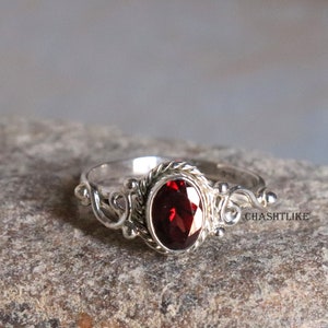 Op de afbeelding: Een zilveren ring met een rode edelsteen gezet in een delicaat, versierd ontwerp. De ring ligt op een grijs, getextureerd oppervlak.