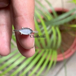 Puede incluir: Un anillo de plata con una gema gris y blanca en forma de marquesa. El anillo tiene una banda delgada y la gema está engastada con cuatro pequeñas garras. El anillo se muestra sobre un fondo de follaje verde.