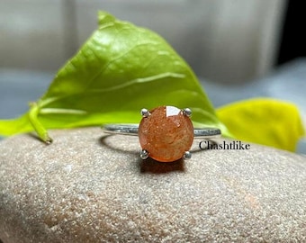 May include: A silver ring with a round, orange sunstone gemstone. The ring is set on a light brown stone.