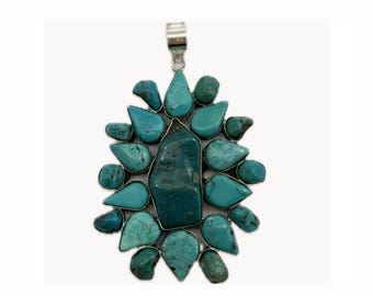 May include: A large turquoise pendant with a silver-colored bail. The pendant features a central, irregularly shaped turquoise stone surrounded by smaller, teardrop-shaped turquoise stones. The stones are set in a silver-colored metal.