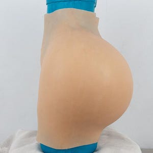 Könnte beinhalten: Ein hautfarbener Silikon-Gesäßverstärker, der die natürliche Form des Trägers vergrößern soll. Die Prothese wird auf einem blauen Ständer präsentiert, der ihre Form und Konturen zeigt. Das Material wirkt glatt und realistisch.