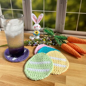 May include: Four crocheted egg-shaped coasters in pastel colors with white stripes. The coasters are on a wooden surface with a glass of water and a ceramic bunny figurine.