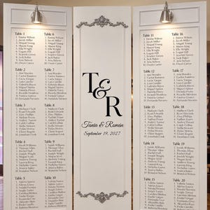 May include: A white three-panel wedding seating chart with silver-toned decorative molding. The center panel features the initials T & R, the names Tania & Román, and the date September 19, 2027. The side panels list tables 1 through 20.