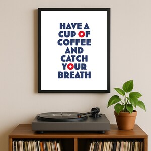 Può includere: Stampa incorniciata con il testo "HAVE A CUP OF COFFEE AND CATCH YOUR BREATH" in blu e rosso. La stampa è esposta sopra un giradischi su un mobile in legno. Una pianta in vaso è a destra.