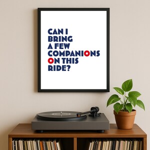 Può includere: Una stampa incorniciata con il testo "CAN I BRING A FEW COMPANIONS ON THIS RIDE?" in blu e rosso. Un giradischi è su un mobile di legno, con una pianta in un vaso di terracotta. Sotto sono visibili dei dischi in vinile.