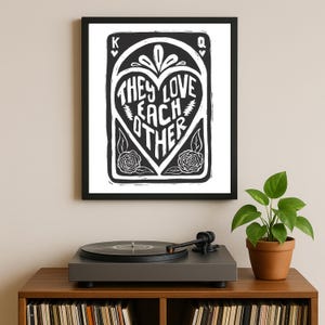 Puede incluir: Impresión artística enmarcada en blanco y negro con el texto "THEY LOVE EACH OTHER" dentro de un diseño en forma de corazón. La impresión se muestra en un mueble de madera con un tocadiscos y una planta en maceta.