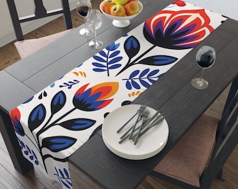 May include: A white table runner with a colorful floral pattern in shades of blue, red, orange, and black. The runner is laid on a dark wood table with two chairs and a bowl of apples.