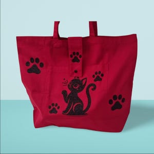 Peut inclure: Sac fourre-tout en toile rouge avec un chat noir pailleté et un motif d'empreintes de pattes. Le sac a une poche sur le devant avec une fermeture à bouton.
