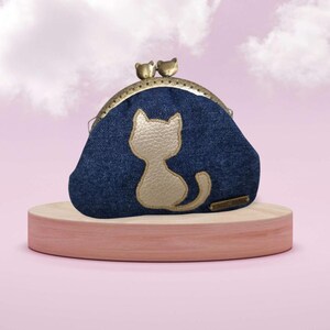 Op de afbeelding: Een denim portemonnee met een gouden kattenmotief. De portemonnee heeft een metalen sluiting met twee kleine katvormige accenten. Het staat op een ronde, lichtgekleurde houten standaard tegen een roze achtergrond.