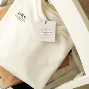 Pode incluir: Uma camisa de moletom branca com o texto "BRIDE" e "not responsible for things said while wedding planning" impresso nela. Uma etiqueta branca com o texto "BLOOM WITH ME DESIGNS" está presa à camisa de moletom.