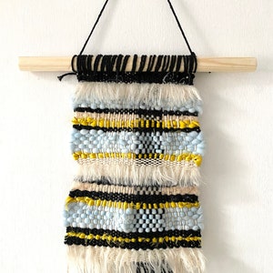 May include: A woven wall hanging with a beige, blue, yellow, and black geometric pattern. The hanging has a wooden dowel at the top and fringe at the bottom.