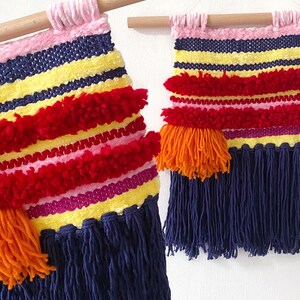 May include: Two colorful woven wall hangings with fringe. The larger hanging has red, pink, yellow, and blue stripes with orange pom-poms. The smaller hanging has the same colors and pattern but is smaller in size.