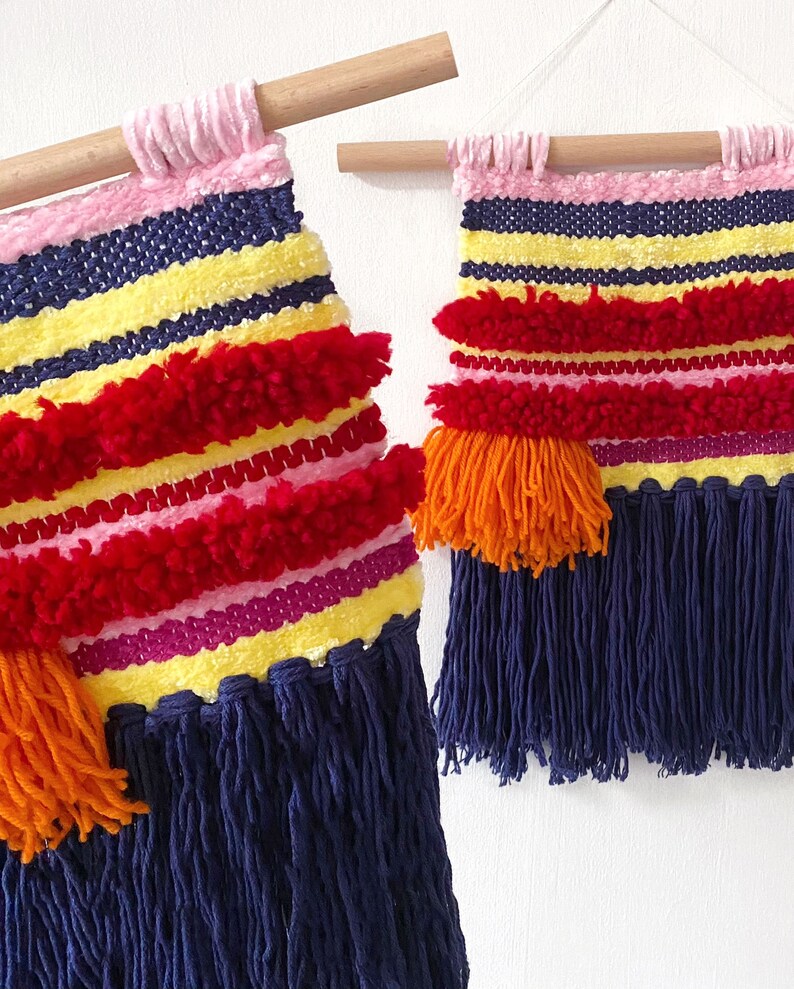 May include: Two colorful woven wall hangings with fringe. The larger hanging has red, pink, yellow, and blue stripes with orange pom-poms. The smaller hanging has the same colors and pattern but is smaller in size.