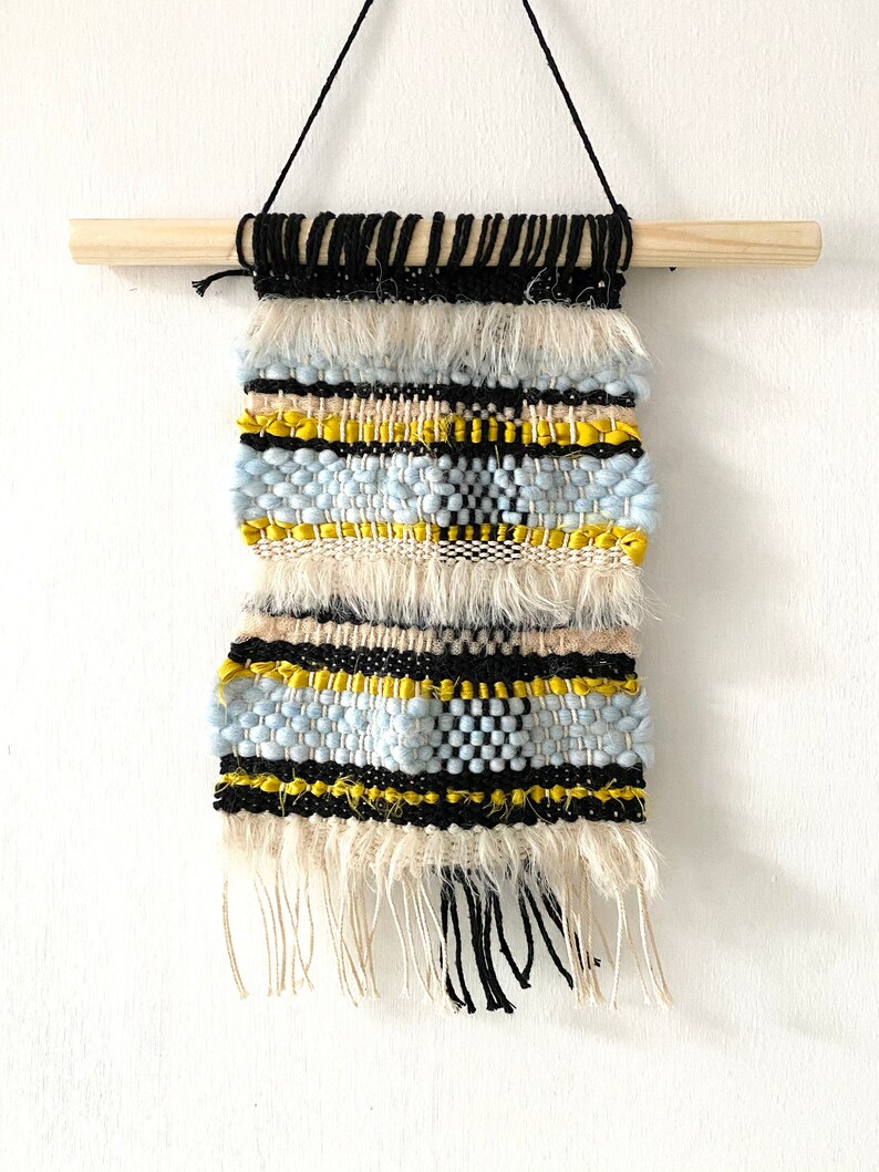 May include: A woven wall hanging with a beige, blue, yellow, and black geometric pattern. The hanging has a wooden dowel at the top and fringe at the bottom.