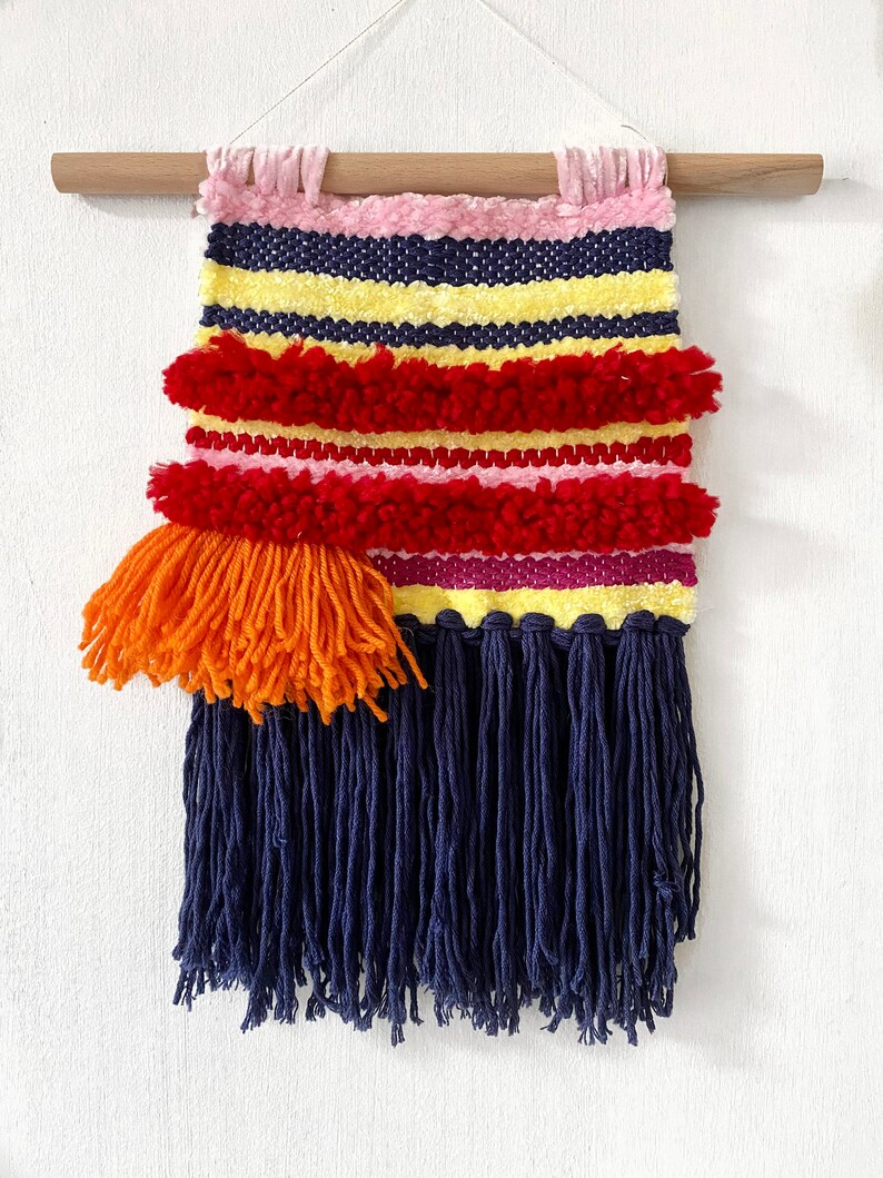 May include: A colorful woven wall hanging with a fringe border. The hanging features stripes of yellow, pink, red, and blue yarn. A large orange pom-pom is attached to the left side of the hanging.