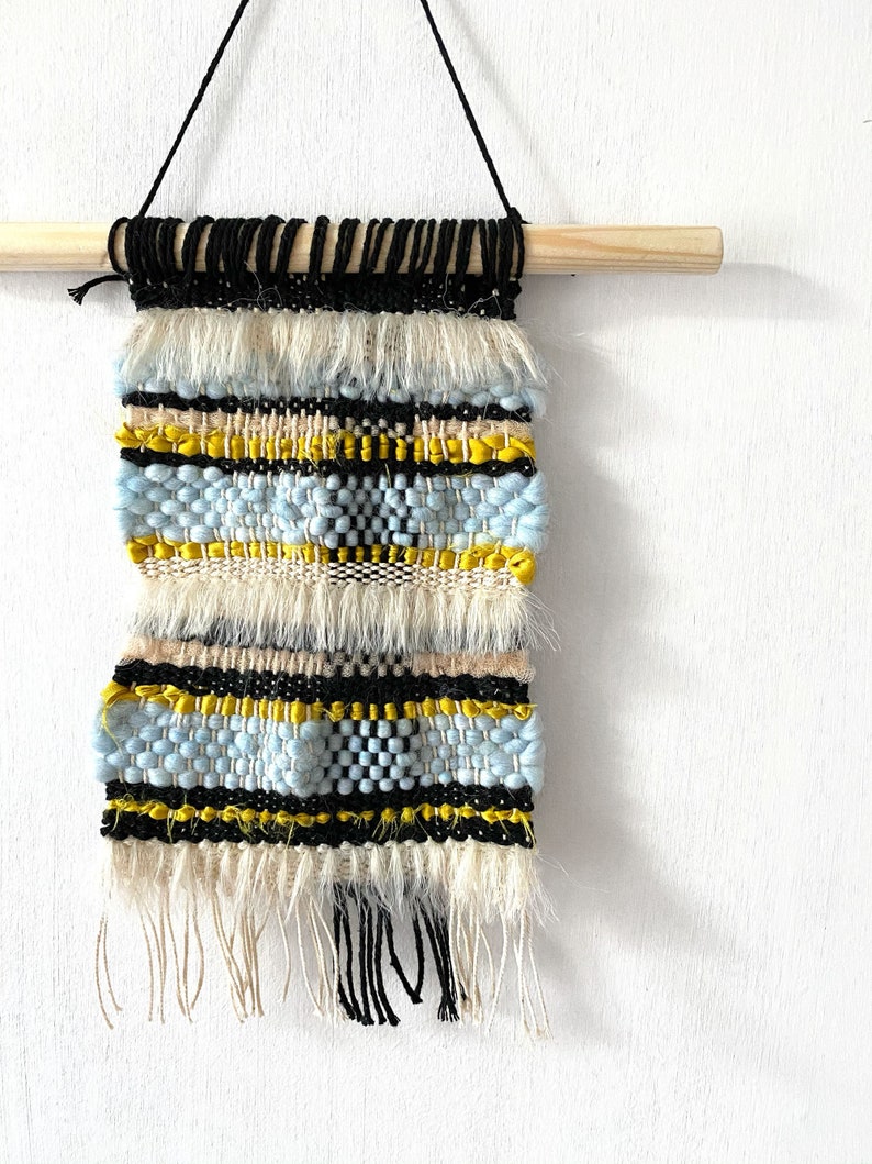 May include: A woven wall hanging with a wooden dowel at the top. The hanging features a pattern of black, white, yellow, and blue yarn. The hanging has fringe at the bottom.