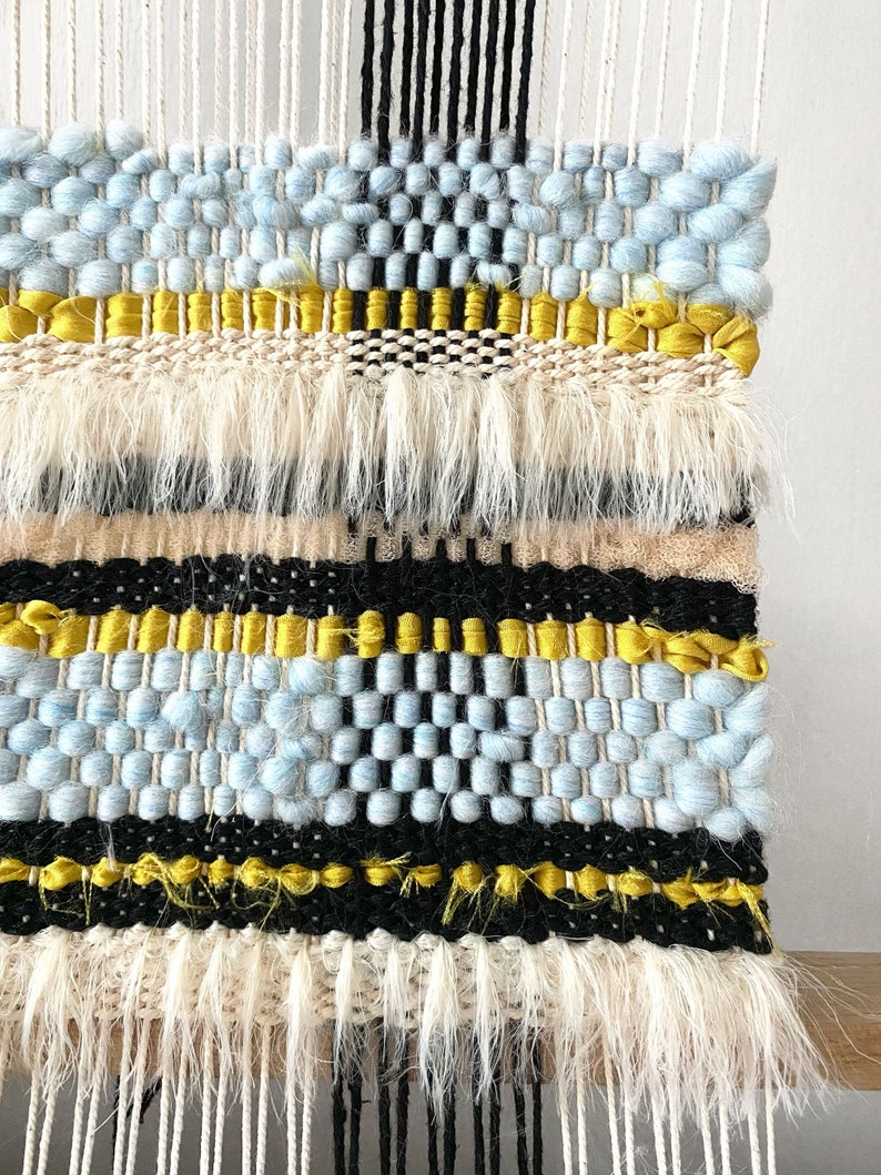 May include: A woven wall hanging with a blue, black, and yellow geometric pattern. The hanging has a fringe border.