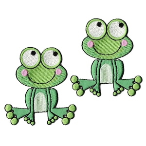 May include: Two embroidered iron-on patches featuring cartoon frogs. Each frog is bright green with large white eyes, black pupils, pink cheeks, and a light green belly. The frogs are depicted in a seated position with splayed legs and toes.