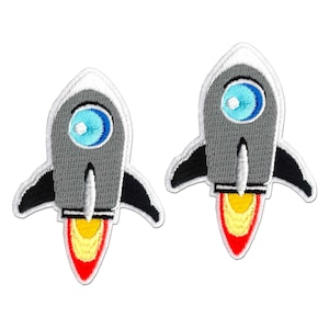 May include: Two embroidered rocket ship patches featuring a gray body, a bright blue circular window, black side fins, and a flame exhaust trail colored in yellow, orange, and red. Each patch has a white border and a detailed stitched texture.