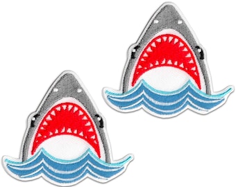 May include: Two embroidered patches depicting a shark's open mouth with red teeth and a grey head. The shark is emerging from blue and white waves. The patches are designed to be ironed on or sewn onto fabric.