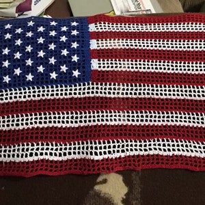 Op de afbeelding: Een gehaakt Amerikaans vlaggenkleed in rood, wit en blauw. De vlag is gemaakt met een gaaspatroon en heeft een gefranst randje.