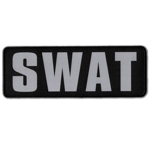 May include: A rectangular black patch featuring the word SWAT in large, bold, light gray capital letters. The patch has a clean, stitched border and a flat, uniform surface, designed for attachment to clothing or gear with a clear, high-contrast design.