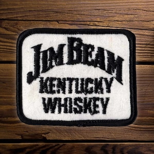 May include: A rectangular embroidered patch with a white background and a thick black border. The patch features the black embroidered text Jim Beam in a stylized font, followed by the words Kentucky Whiskey in a bold, black sans-serif font.