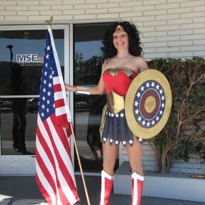 Op de afbeelding: Een vrouw verkleed als Wonder Woman, met een rood, blauw en gouden kostuum, die een schild met sterren vasthoudt en een rode ster op haar voorhoofd. Ze staat voor een gebouw met een vlaggenmast en een Amerikaanse vlag.