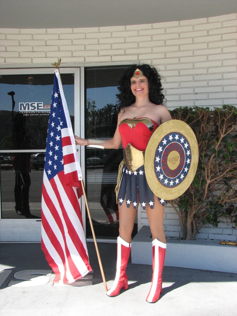 Op de afbeelding: Een vrouw verkleed als Wonder Woman, met een rood, blauw en gouden kostuum, die een schild met sterren vasthoudt en een rode ster op haar voorhoofd. Ze staat voor een gebouw met een vlaggenmast en een Amerikaanse vlag.