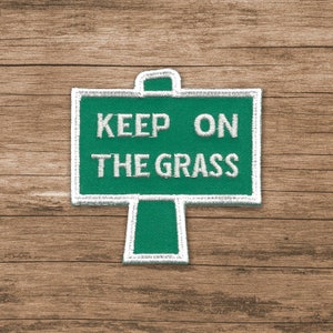May include: A green and white embroidered patch with the text "Keep on the grass" on a wooden background.