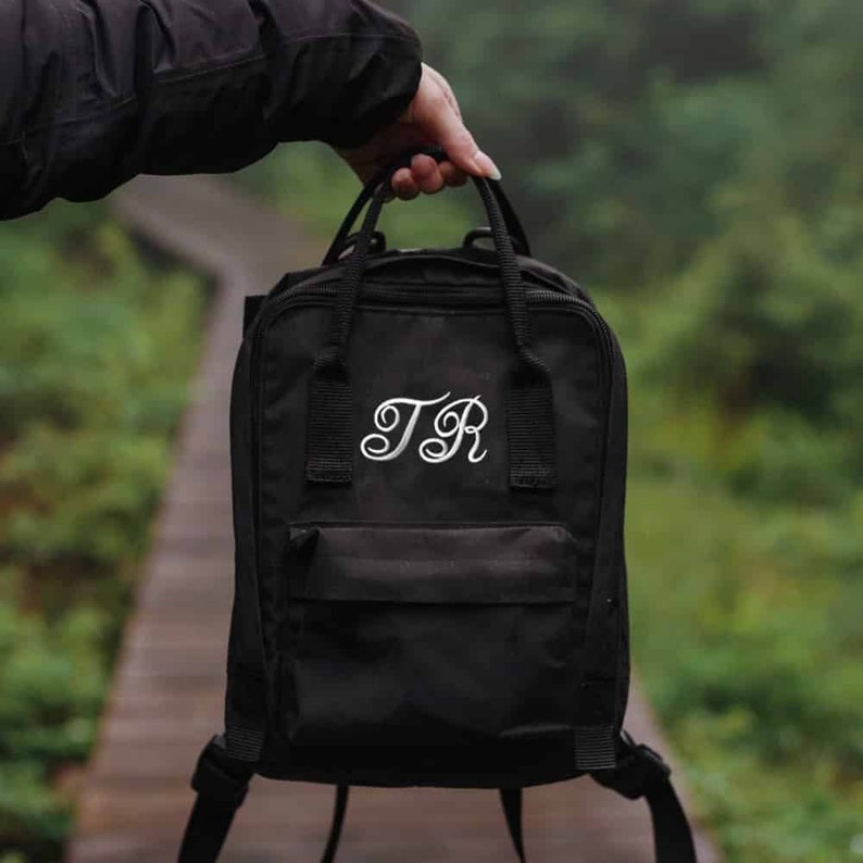 K&ouml;nnte beinhalten: Ein schwarzer Rucksack mit einem wei&szlig;en gestickten Monogramm "TR" auf der Vorderseite.