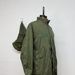 Op de afbeelding: Olijfgroene jas in militaire stijl met een hoge kraag en meerdere zakken. De jas wordt op een mannequin getoond en heeft een bijpassende capuchon in de buurt hangen. De jas lijkt gemaakt van een duurzaam, mogelijk waterafstotend materiaal.