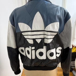 May include: A vintage leather jacket with a color block design. The jacket features a blue, white, black, and gray color scheme with the Adidas logo and brand name prominently displayed on the back. The jacket has a black collar and cuffs.