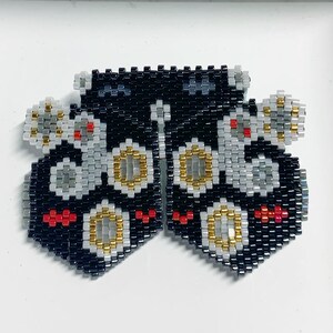 May include: Handmade beaded earrings in a butterfly shape. The earrings feature a black background with gray, gold, and red accents. The intricate beadwork creates a geometric pattern, showcasing the craftsmanship of the jewelry.