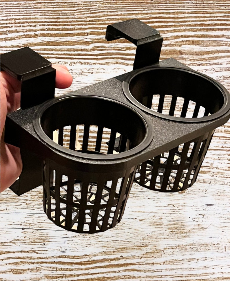 May include: Black plastic double cup holder with a slotted basket design. The holder has a hanging bracket and is designed to hold two cups or containers. The cup holder is shown against a light wood background.