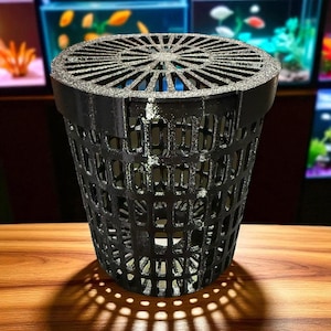 May include: Black plastic hydroponic net pot with a slotted design for aquatic plant cultivation. The pot has a circular top with radial slots and a cylindrical body with rectangular openings. The pot is sitting on a wooden surface.