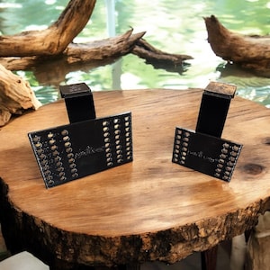 Puede incluir: Dos tarjeteros de metal negro con un diseño perforado y un pequeño soporte. Los tarjeteros se exhiben sobre una mesa de madera con borde natural. El fondo presenta agua y madera flotante.