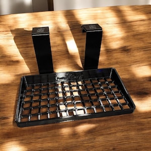 Puede incluir: Una bandeja de teclado de metal negro con dos soportes verticales. La bandeja tiene un patrón de rejilla y está diseñada para fijarse a un escritorio. La bandeja se muestra sobre una superficie de madera.