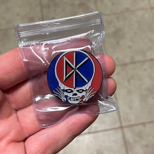 May include: A red, white, and blue enamel pin with a skull and crossbones design. The skull is wearing a blue and white hat with a red band. The pin is in a clear plastic bag.
