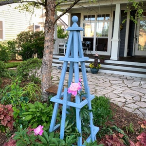 May include: A light blue wooden garden obelisk with a ball finial, supporting pink peony flowers. The obelisk is set in a garden with lush green foliage and a stone patio in the background.