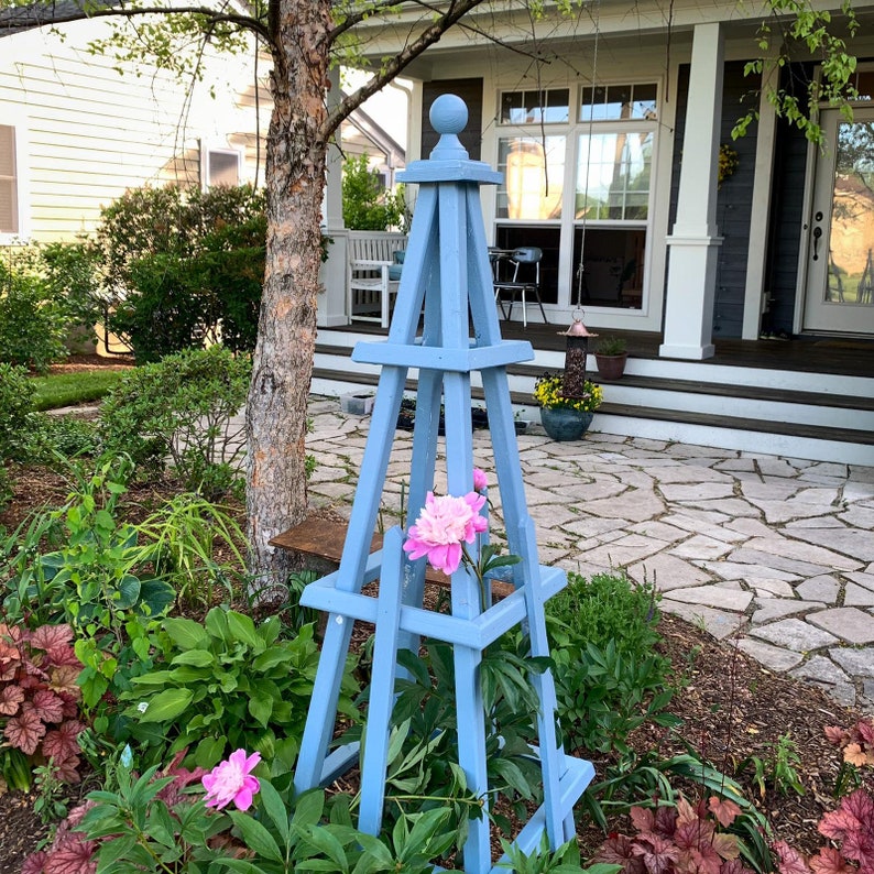 May include: A light blue wooden garden obelisk with a ball finial, supporting pink peony flowers. The obelisk is set in a garden with lush green foliage and a stone patio in the background.