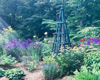 May include: A lush garden scene featuring a blue wooden obelisk surrounded by vibrant flowers. Purple, white, and yellow blooms are interspersed among green foliage. The obelisk is a decorative garden element.