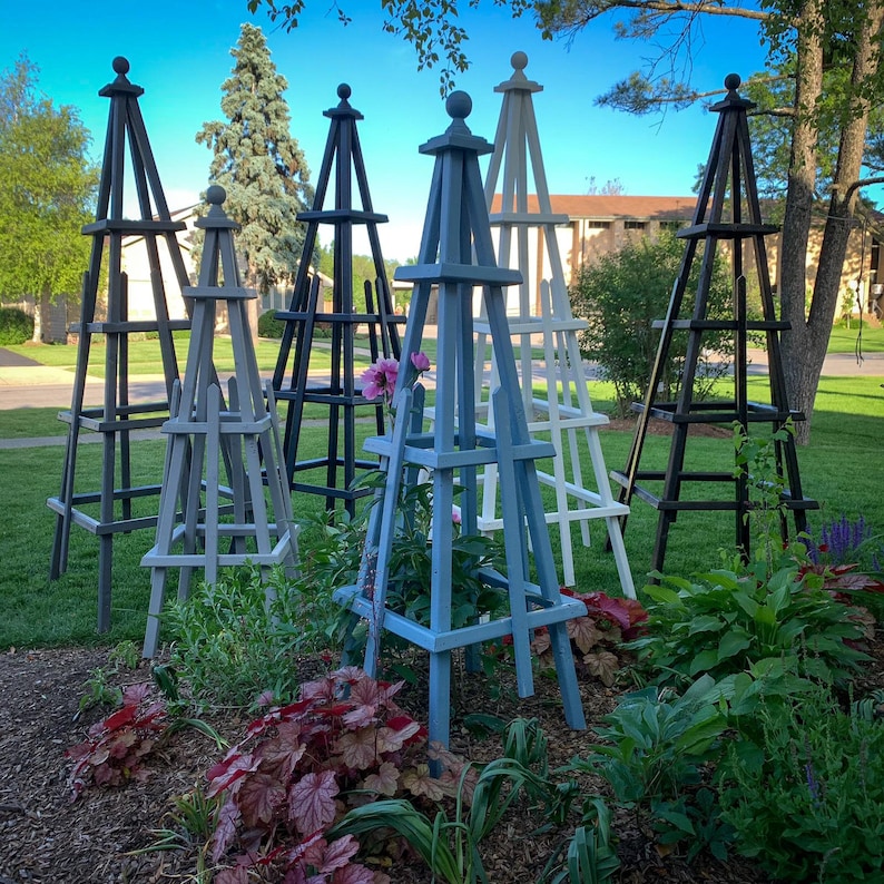 May include: A collection of wooden garden obelisks in various colors, including black, gray, white, and blue. The obelisks are pyramid-shaped with a decorative finial at the top, and are placed in a garden bed with plants.