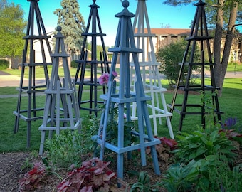 May include: A collection of wooden garden obelisks in various colors, including black, gray, white, and blue. The obelisks are pyramid-shaped with a decorative finial at the top, and are placed in a garden bed with plants.