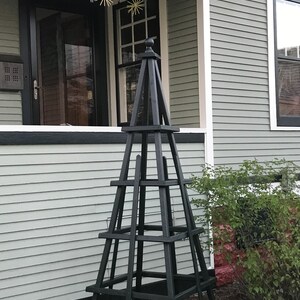 May include: A tall, black wooden obelisk-style garden structure with a square base and multiple horizontal supports. The structure is placed in front of a house with light green siding and a black door and window trim. Decorative star-shaped ornaments hang from the porch.