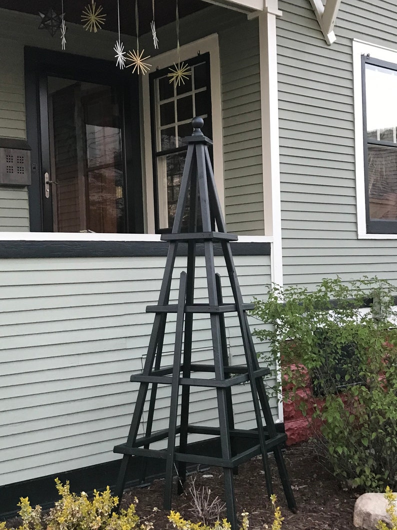 May include: A tall, black wooden obelisk-style garden structure with a square base and multiple horizontal supports. The structure is placed in front of a house with light green siding and a black door and window trim. Decorative star-shaped ornaments hang from the porch.