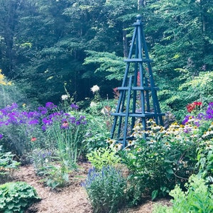 May include: A vibrant garden scene featuring a teal obelisk amidst a profusion of colorful flowers. Purple, white, yellow, and red blooms create a lively display. Lush green foliage and trees form a natural backdrop, bathed in sunlight.