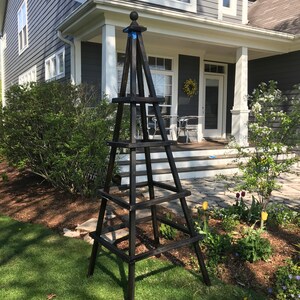 May include: A tall, dark brown wooden garden obelisk with a ball finial. The obelisk has a pyramid shape with horizontal supports. It stands on a green lawn in front of a house with a gray exterior and white trim. The obelisk is a decorative garden structure.