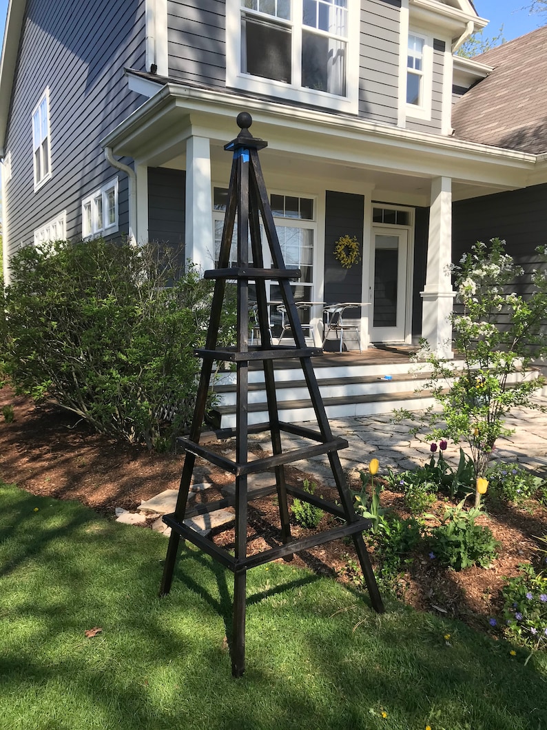May include: A tall, dark brown wooden garden obelisk with a ball finial. The obelisk has a pyramid shape with horizontal supports. It stands on a green lawn in front of a house with a gray exterior and white trim. The obelisk is a decorative garden structure.