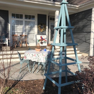 May include: A light blue wooden obelisk stands in a garden setting. The obelisk has a pyramid shape with a square base and is painted in a muted blue color. A small table and chairs are visible in the background.